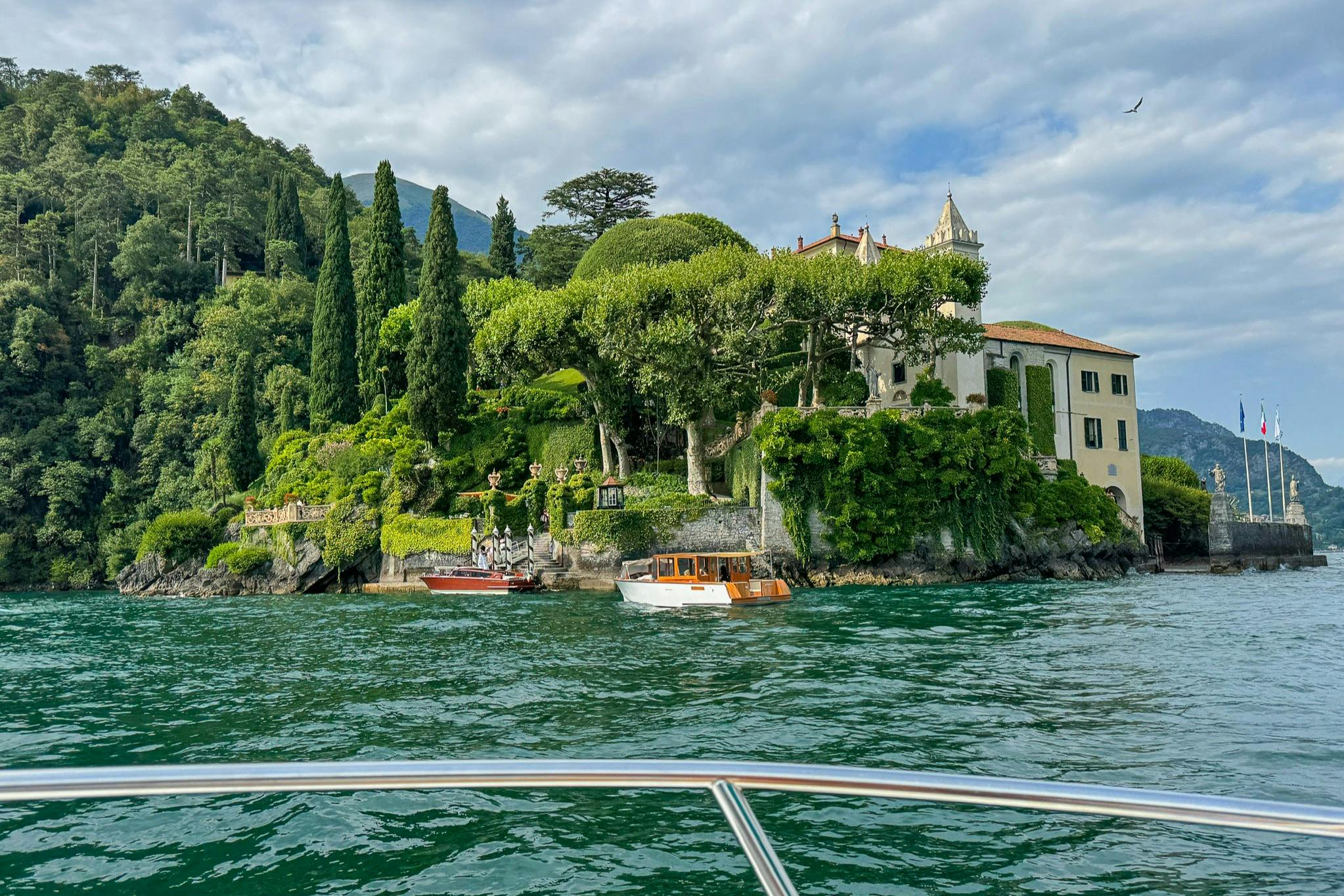 A scenic lakeside view with boats docked near lush greenery and a villa surrounded by trees and a cloudy sky.