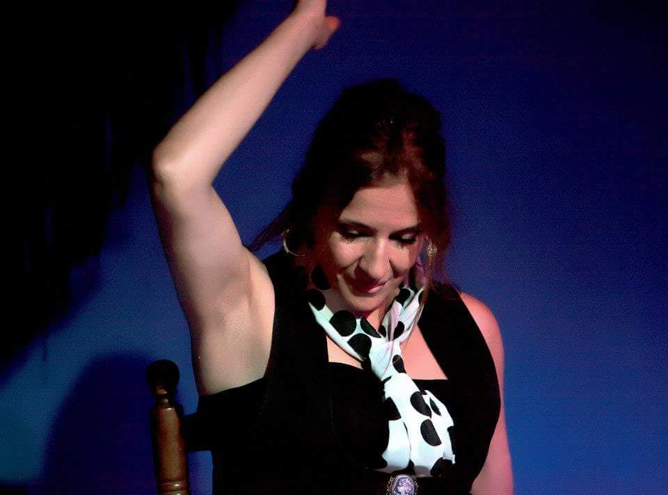 A person wearing a black vest and a white scarf with black dots, seated, with one arm raised, looking downward against a blue background.
