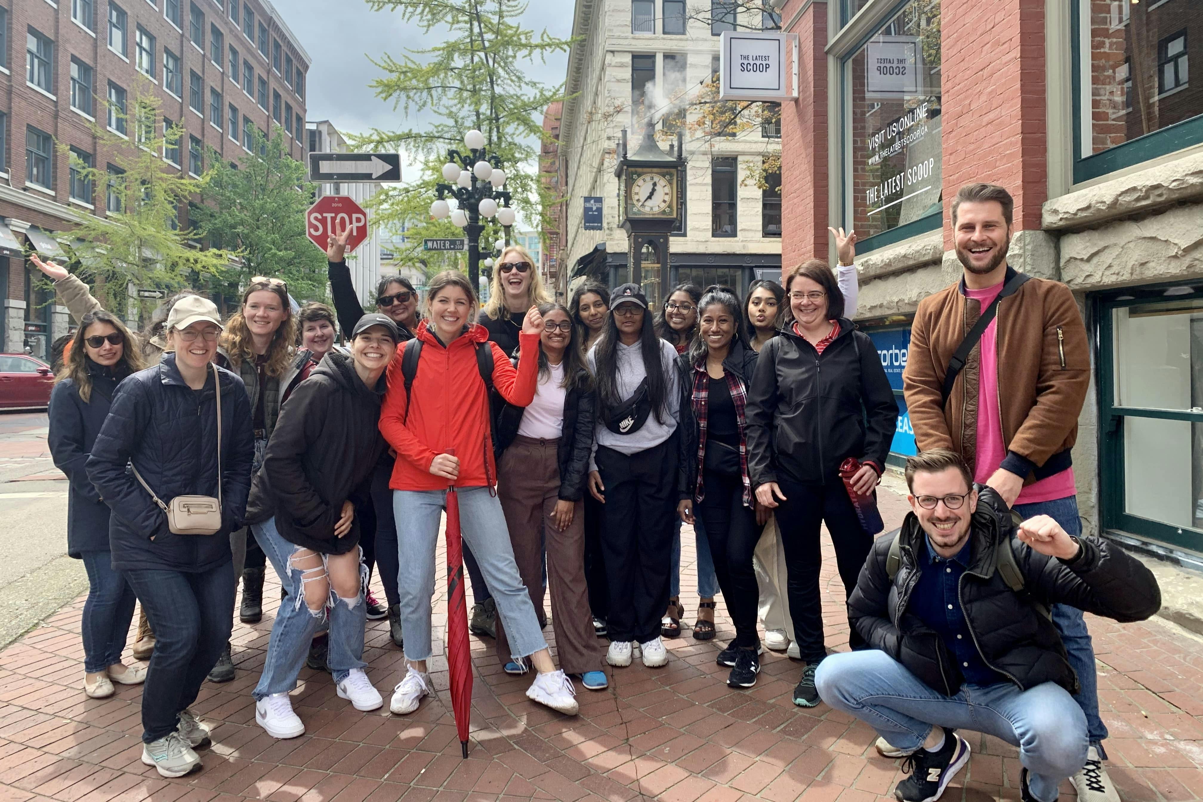 Skupina lidí pózující pro fotografii na dlážděné ulici poblíž parních hodin a značky stop v městské oblasti.
