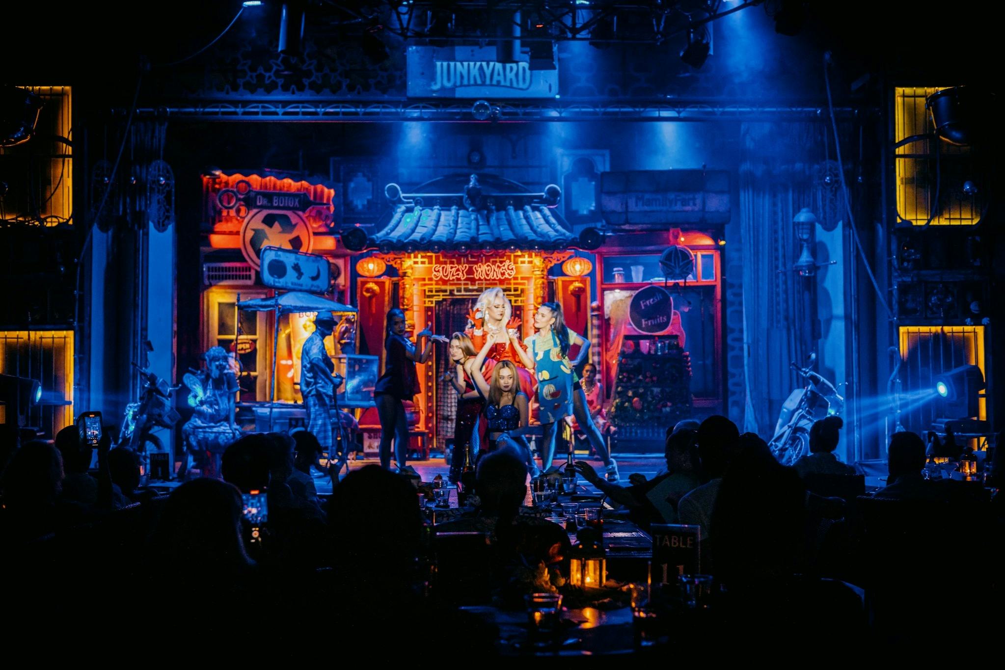 Танцоры выступают на яркой неоновой сцене с аудиторией, наблюдающей на переднем плане. На сценическом знаке написано «Свалка».