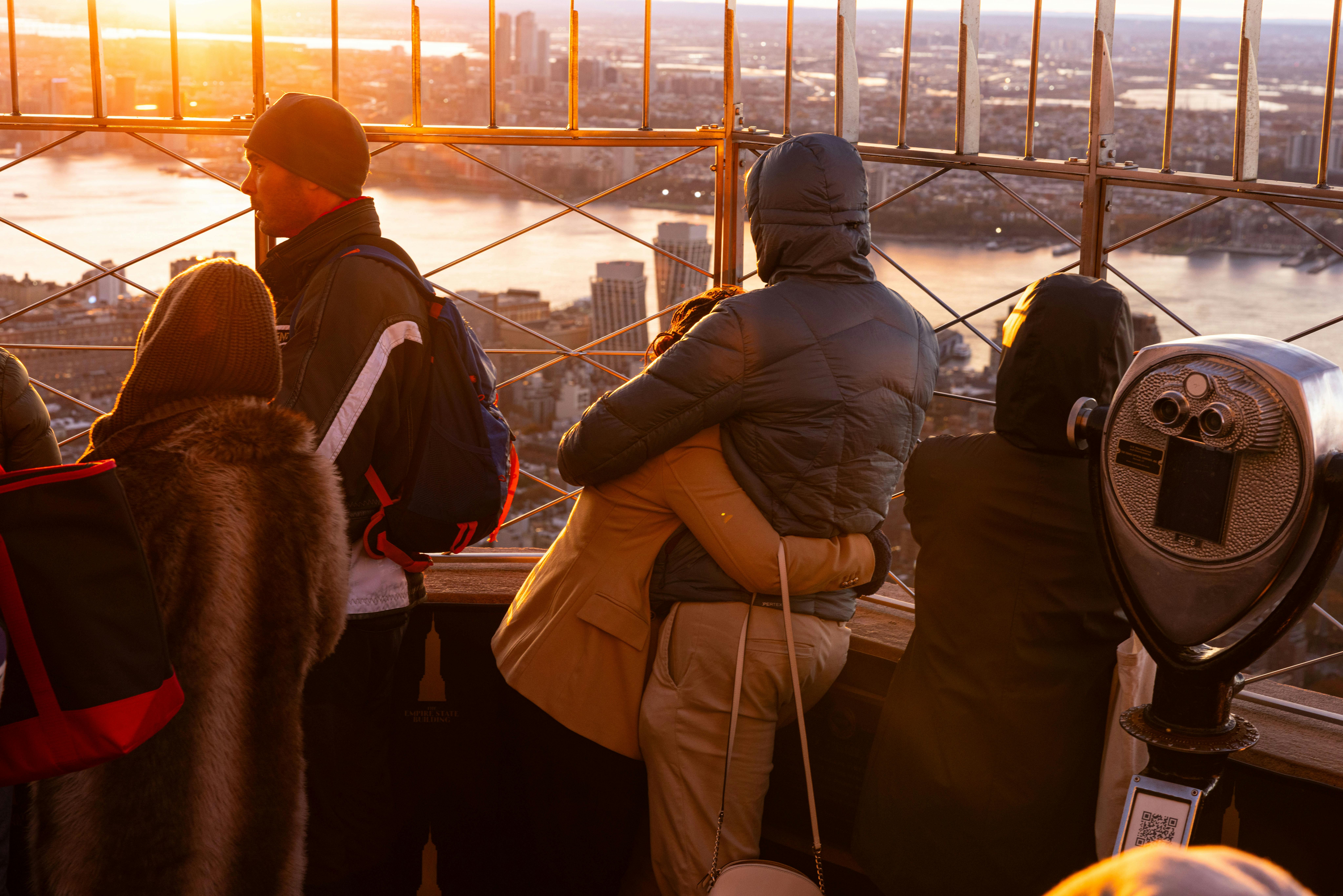People on an observation deck at sunset overlooking a cityscape and a river, with warm sunlight illuminating the scene.