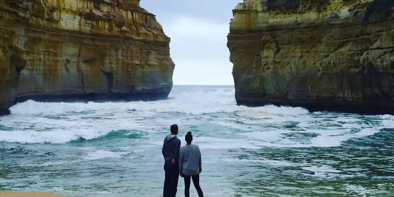 A couple stands on a sandy beach, facing large ocean waves and dramatic rocky cliffs under a cloudy sky.