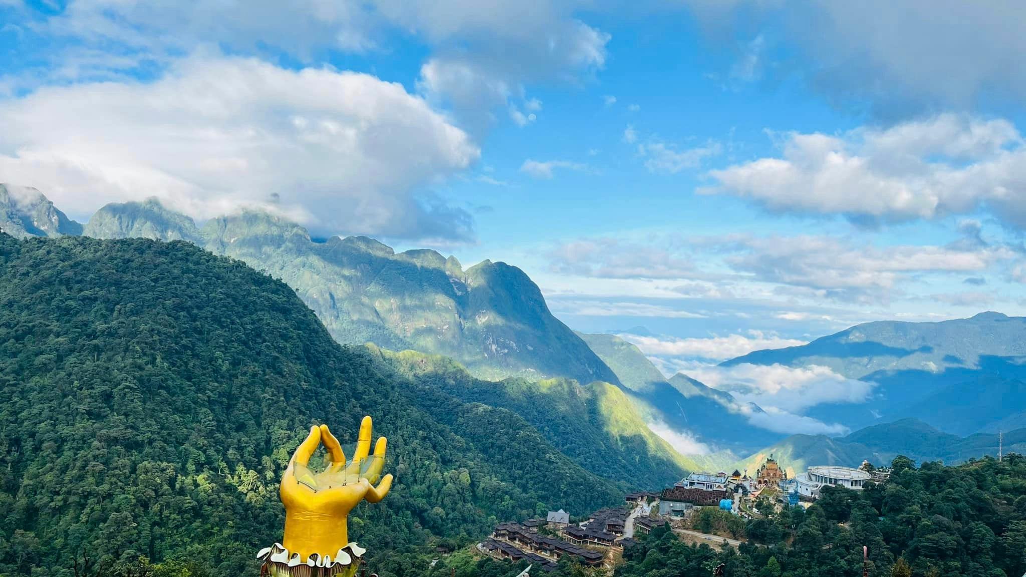 A scenic view of a mountainous landscape under a blue sky with clouds, a yellow hand sculpture, and a village below.