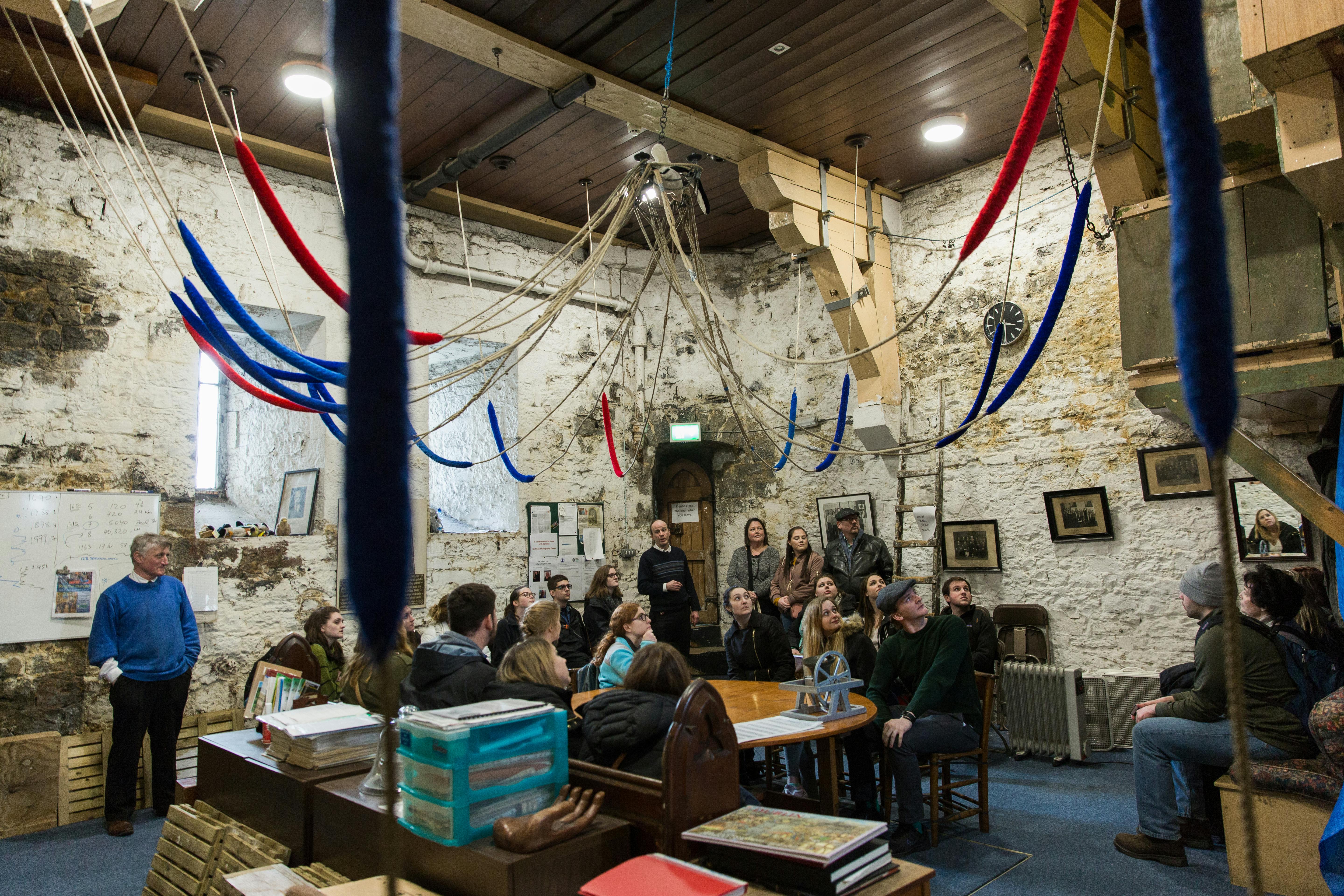 Un gruppo di persone siede e sta in piedi in una stanza rustica con pareti in pietra, corde colorate appese al soffitto e varie immagini incorniciate.