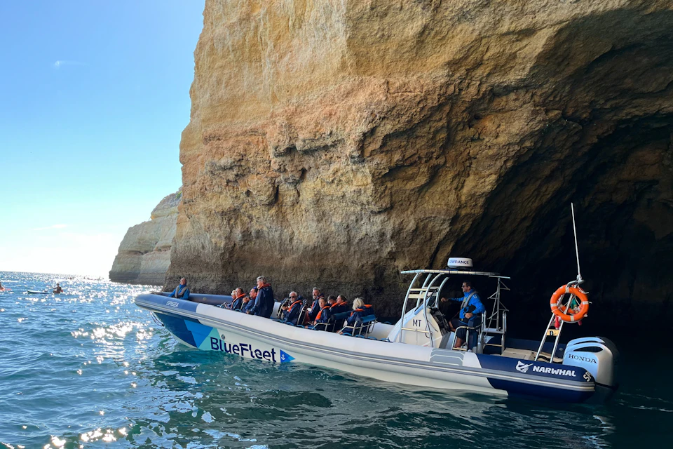 A boat named BlueFleet with passengers wearing life jackets near a rocky coastal cave on a sunny day.