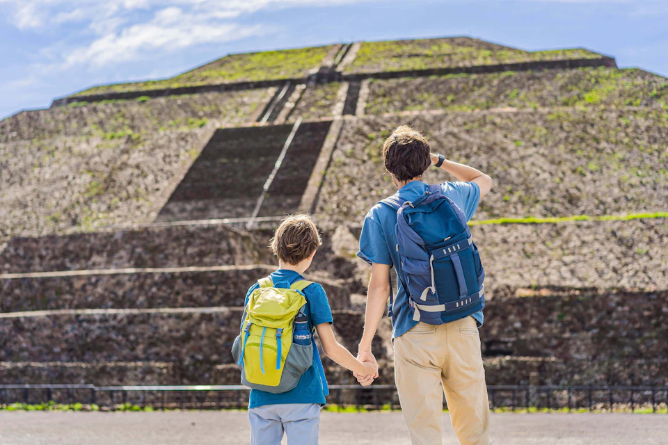 Two people with backpacks hold hands and look at a large ancient stone structure with steps under a clear sky.