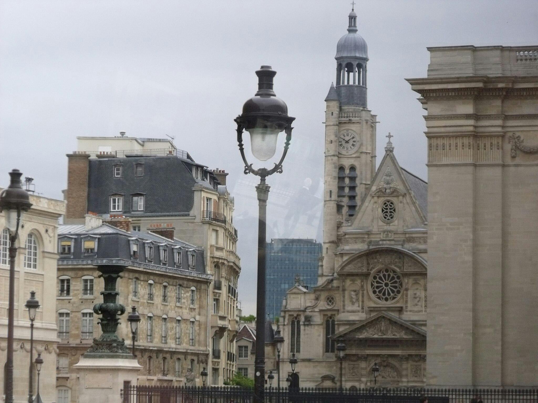 Historisk europæisk gade med udsmykkede bygninger, herunder et klokketårn og vintage gadelamper under en overskyet himmel.
