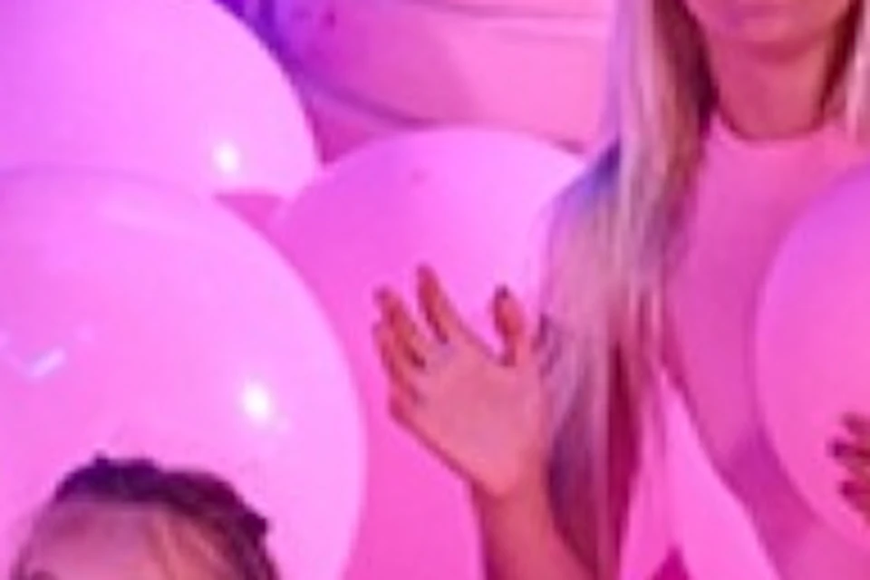 A woman and two girls in a room filled with pink balloons, near a column topped with a large illuminated flower.