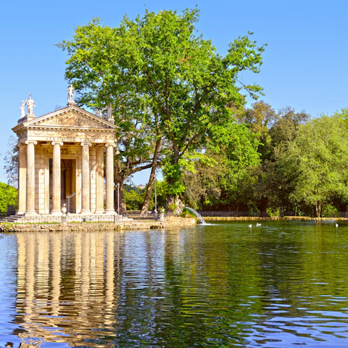 Ett klassiskt tempel med pelare och statyer står vid en fridfull sjö, omgiven av frodiga träd och grönska under en klarblå himmel.