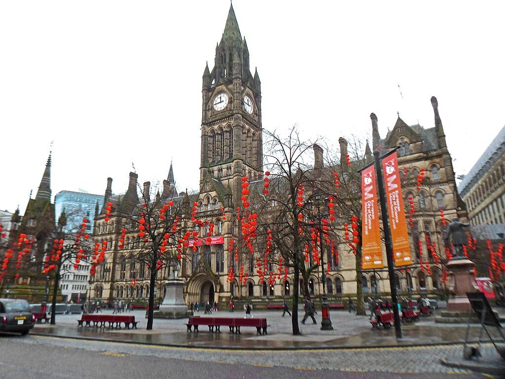 Gothic-style building with a tall clock tower, trees decorated with red lanterns, and banners in a public square.