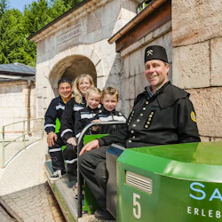 En familj och en guide i gruvuniformer sitter på ett litet tåg utanför en stenbyggnad märkt "FERDINAND.BERG." med "Salz Erlebnisberg" på tåget.