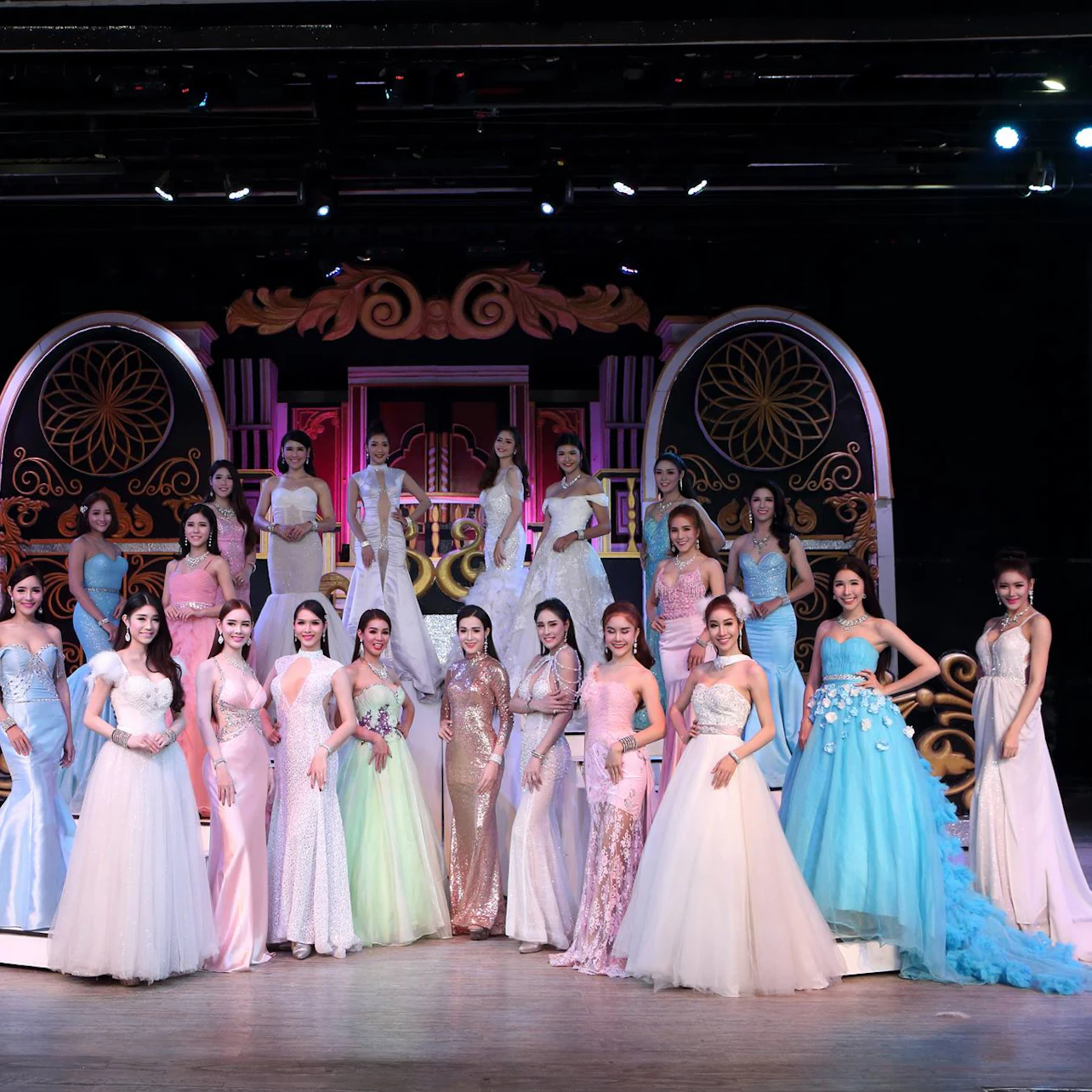 A group of women in elegant gowns pose on a stage with ornate golden and pink decorations.