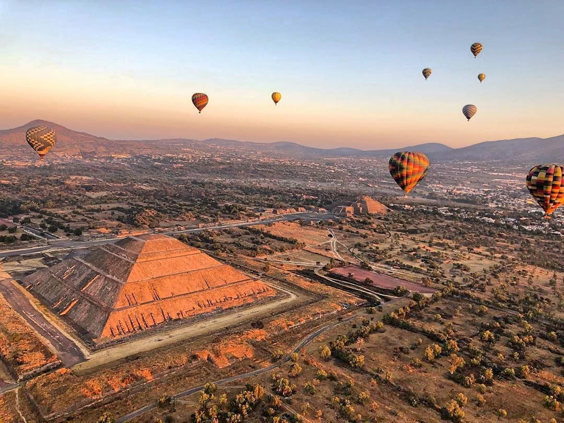 Hot air balloons float over a stepped pyramid and a sprawling landscape at sunrise or sunset, with distant mountains in the background.