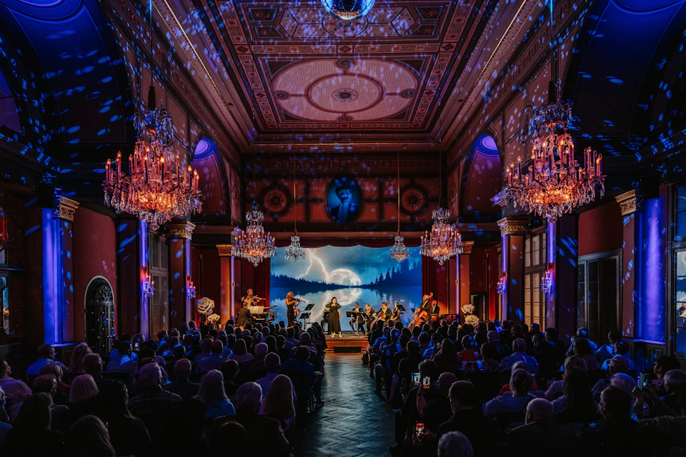 A musical performance in an ornate hall with chandeliers and a backdrop of a moonlit forest scene.