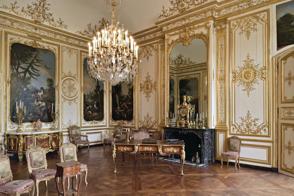 Ornate room with a large chandelier, antique furniture, gilded mirrors, and decorative wall paintings.