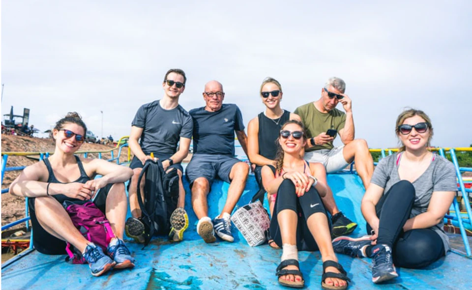Six people sitting on a blue surface outdoors, smiling and wearing casual clothing and sunglasses.