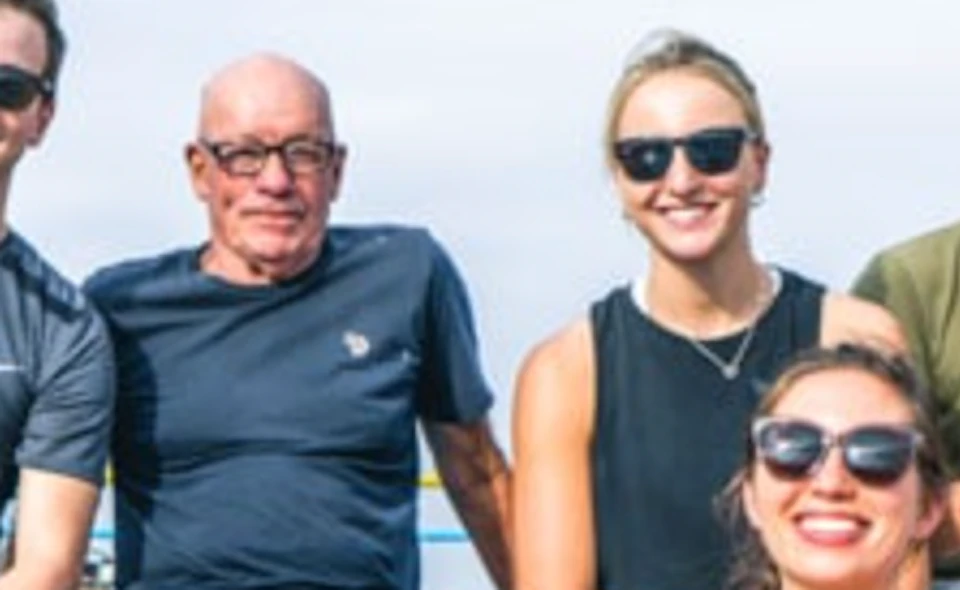 Six people sitting on a blue surface outdoors, smiling and wearing casual clothing and sunglasses.