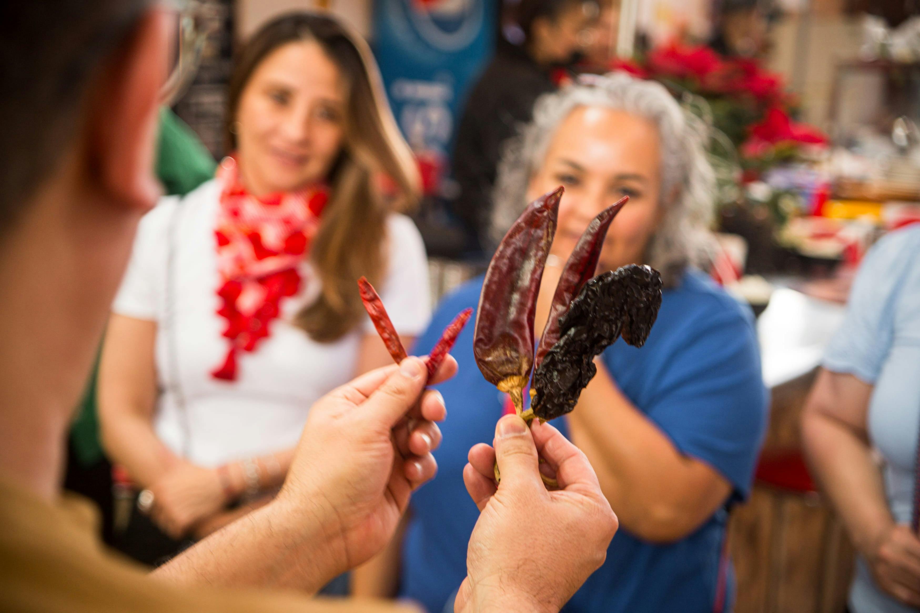 Dos personas observan cómo una persona sostiene varios chiles secos, incluidas variedades rojas y oscuras, en un mercado.