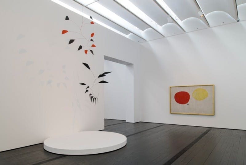 흰색 원형 플랫폼, 나뭇잎 벽화, 빨간색과 노란색 액자에 담긴 예술 작품이 있는 미니멀리즘 갤러리 공간입니다.