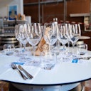 A round table set with multiple wine glasses, water glasses, napkins, cutlery, and paper bags in a restaurant or tasting room.