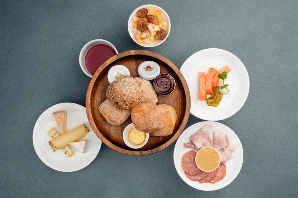 A wooden bowl with bread rolls, butter, and jam, surrounded by plates of cheese, cold cuts, fruit, and salmon sashimi.