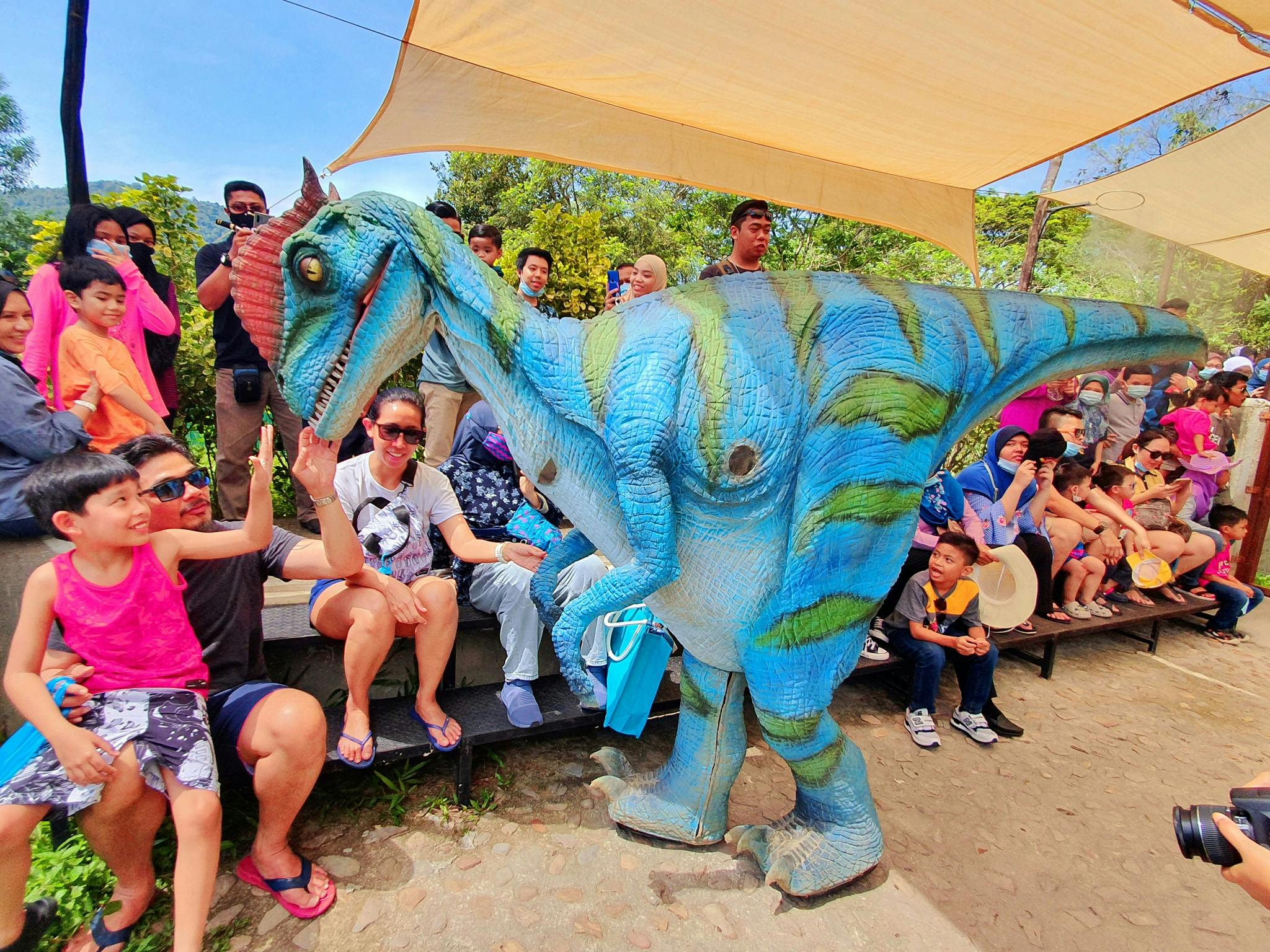 一位穿着恐龙服装的演艺人员在户外与一群微笑的人互动，一些孩子伸出手来触摸它。