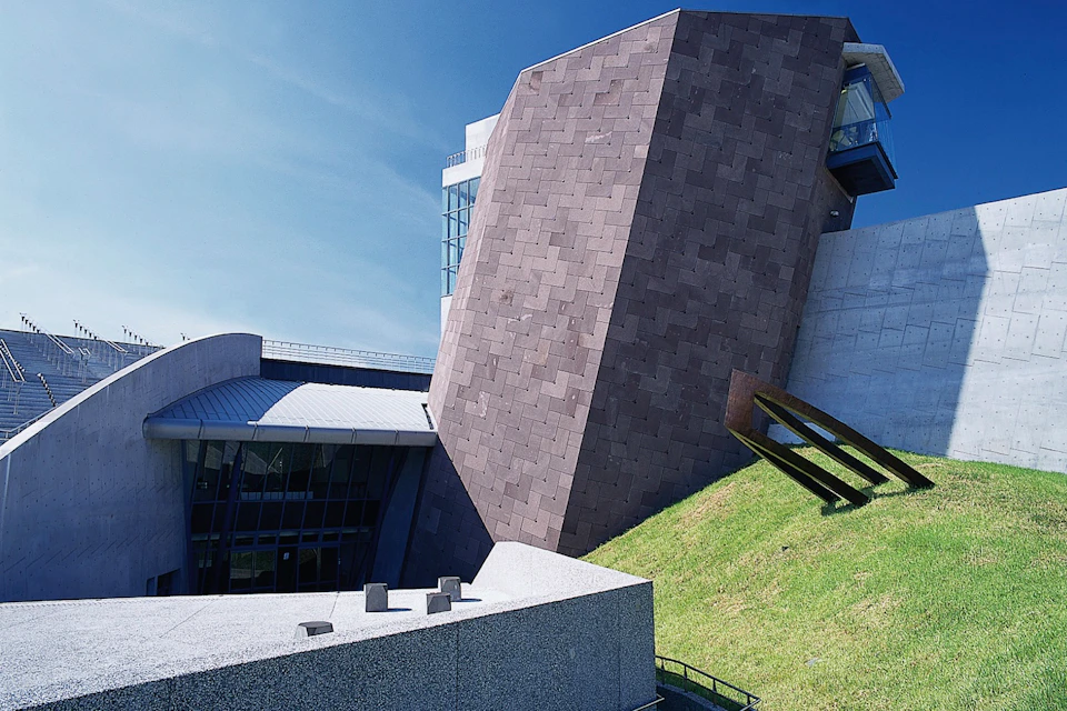 A modern architectural building with angular structures, a grassy slope, and a blue sky in the background.
