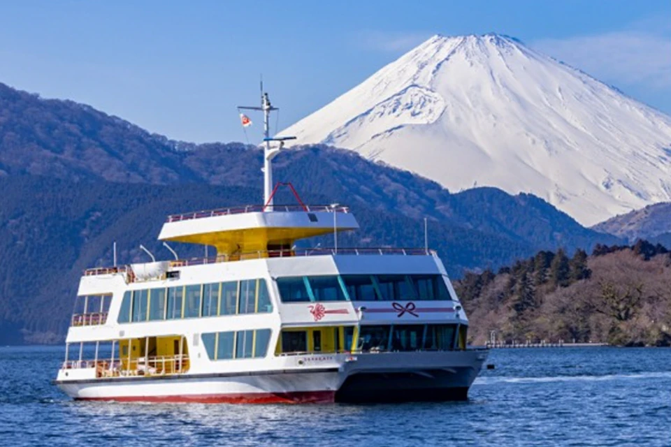 A white ferry with yellow accents on a lake with Mount Fuji in the background under a clear blue sky.