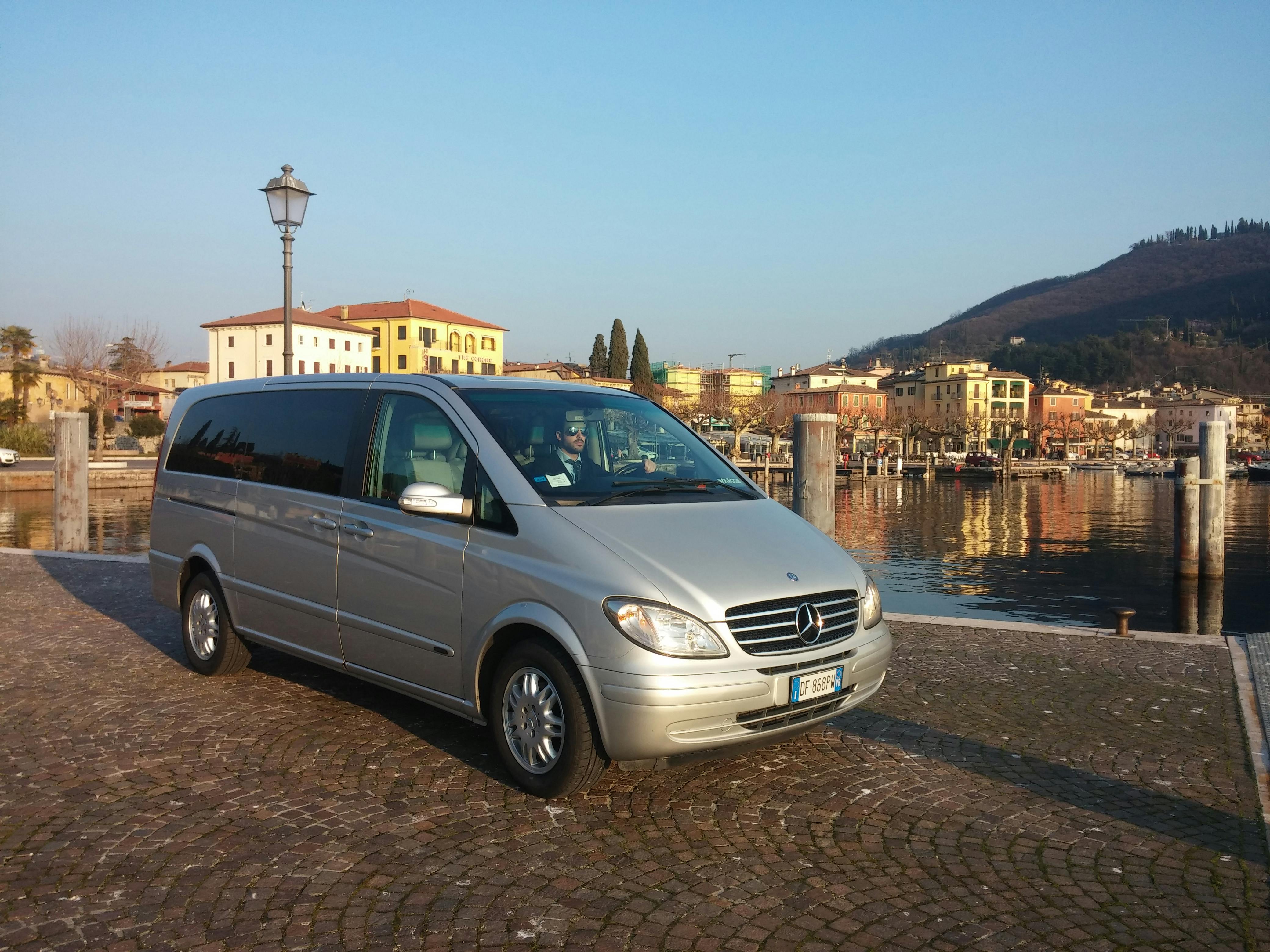 En sølvvarevogn parkeret på en brostensbelagt overflade nær en havnefront med farverige bygninger og bakker i baggrunden.