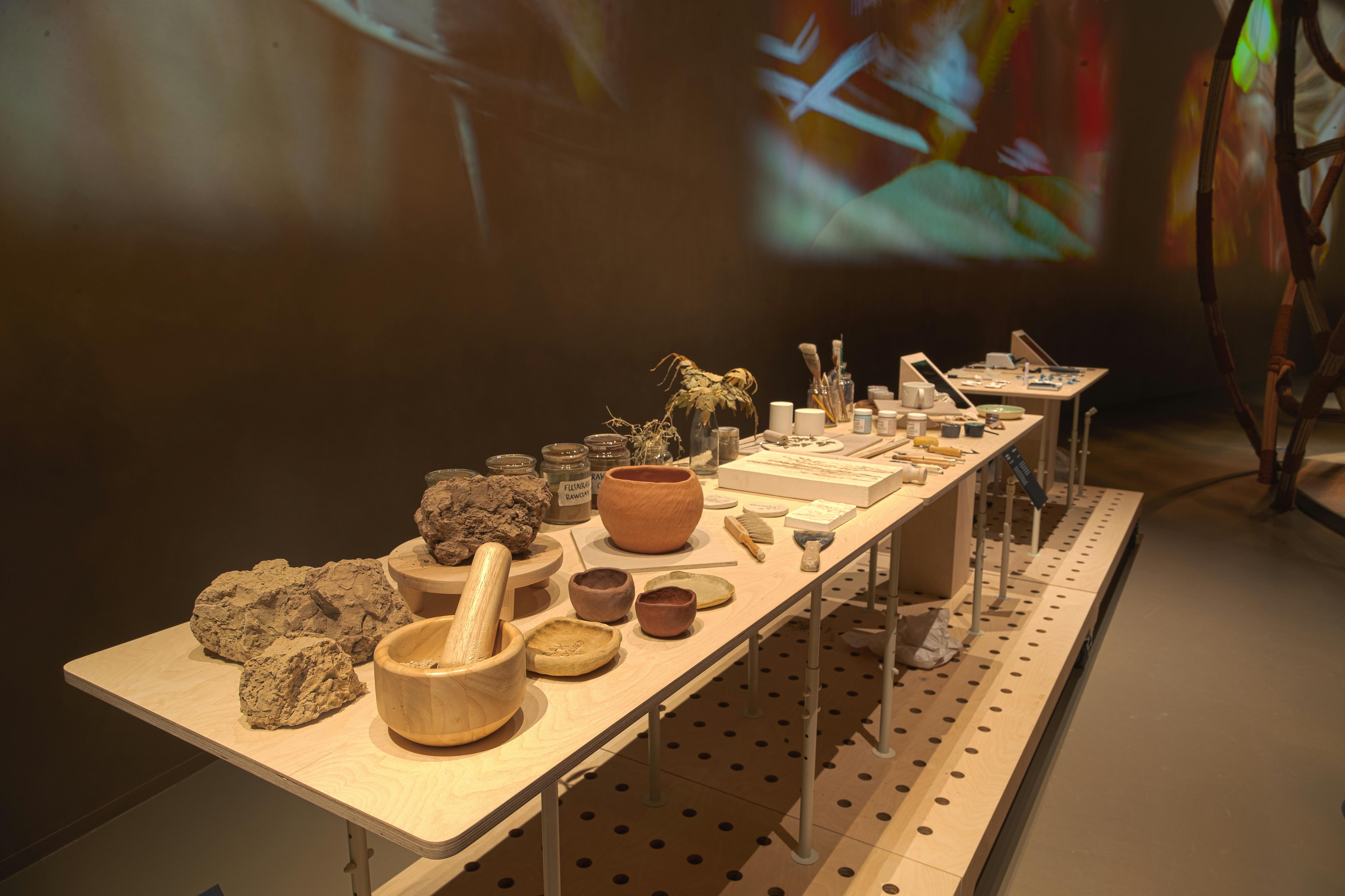 A display of various pottery items, tools, jars, and rocks arranged on a long table against a dark wall with colorful projections.