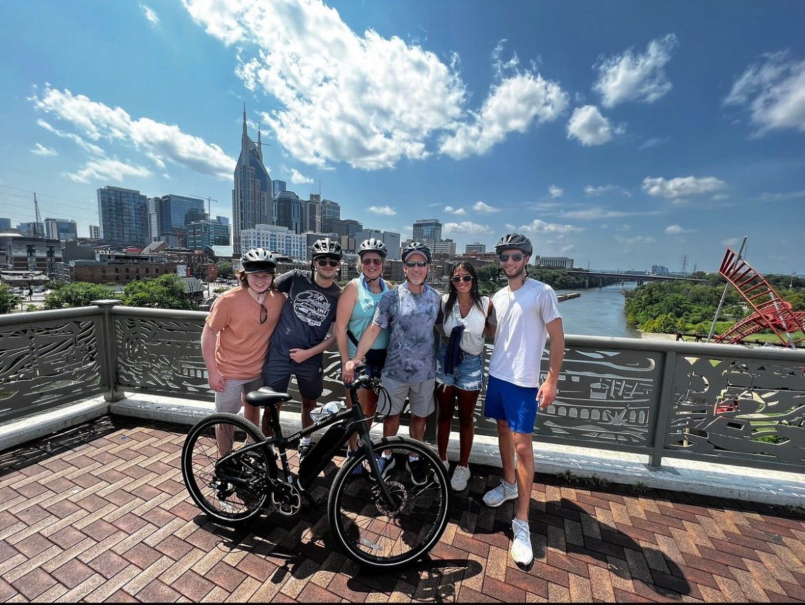 Un grupo de seis personas con bicicletas en un puente, con el horizonte de una ciudad y un río visibles al fondo bajo un cielo soleado.