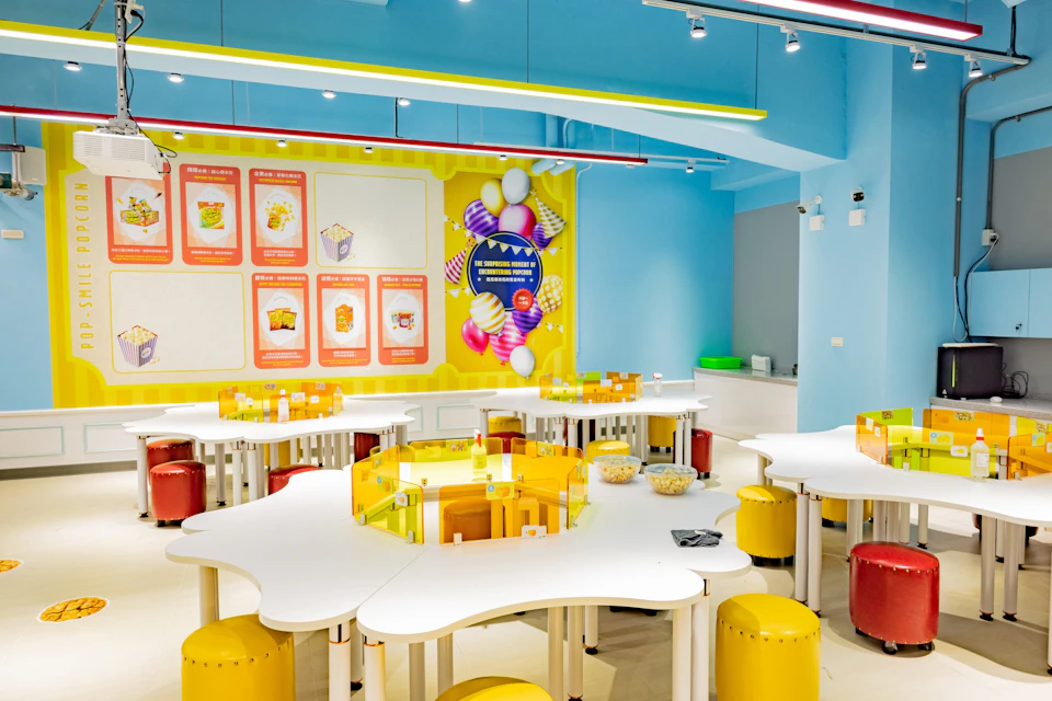 A colorful classroom setup with white tables, yellow stools, and a decorated wall featuring educational displays and illustrations.