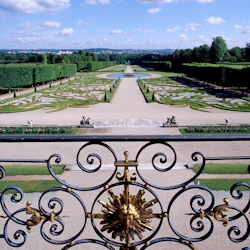 Vy över formella trädgårdar med invecklade häckar och gångar; dekorativt smidesjärnsräcke i förgrunden.