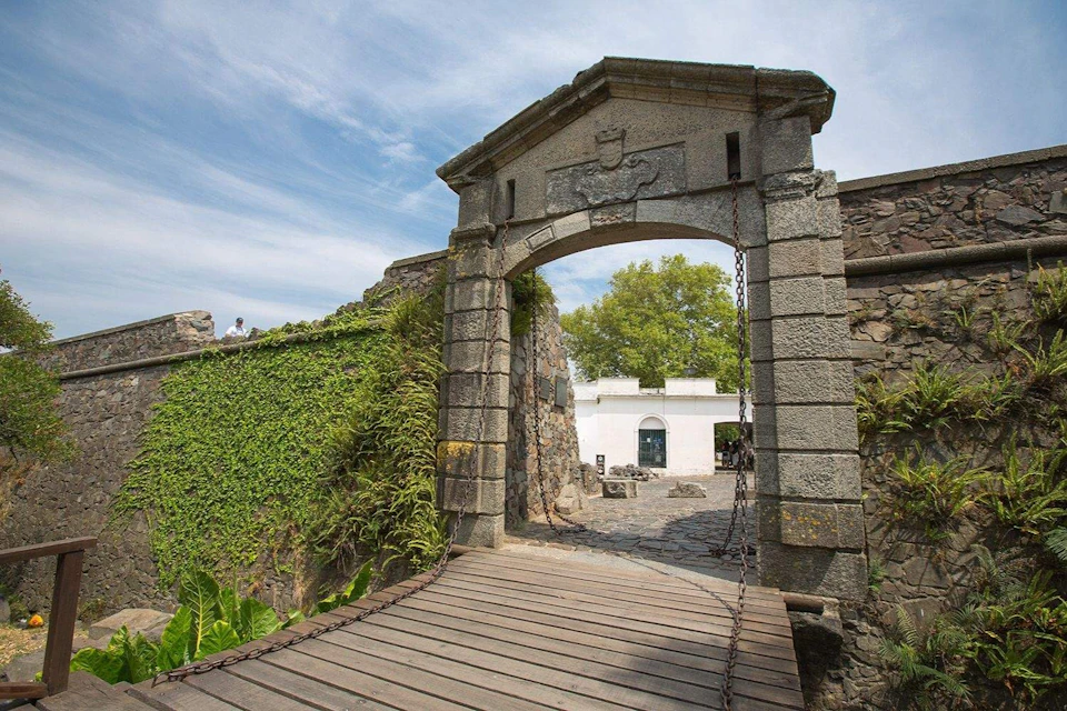 Stone archway with a wooden bridge leading to a courtyard with plants and a white building in the background.