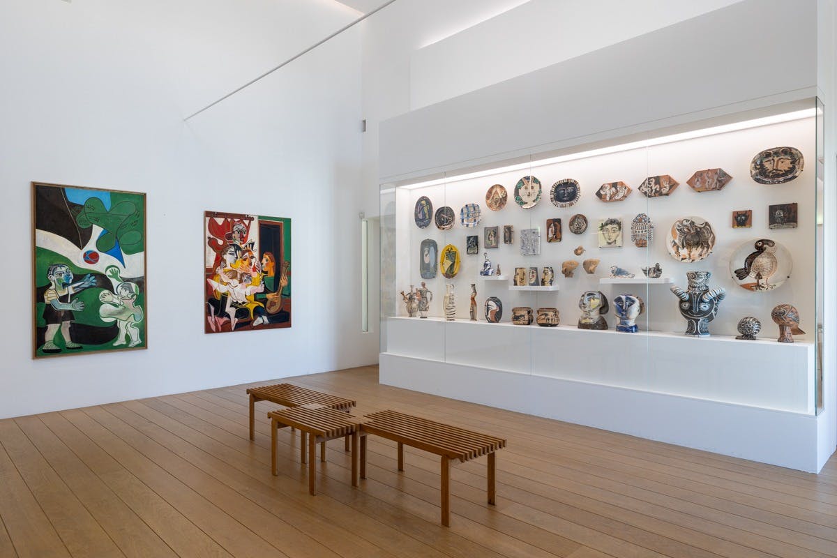 A modern gallery with white walls displaying ceramic artworks in a glass case and paintings on the wall, with wooden benches.