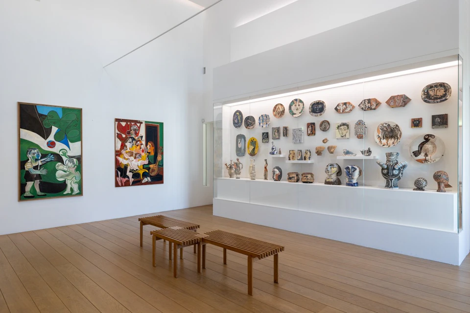 A modern gallery with white walls displaying ceramic artworks in a glass case and paintings on the wall, with wooden benches.