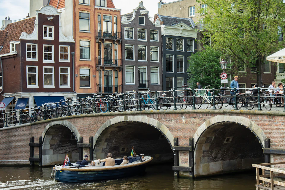 Boat with passengers under a brick bridge lined with bicycles; colorful buildings and trees in the background.