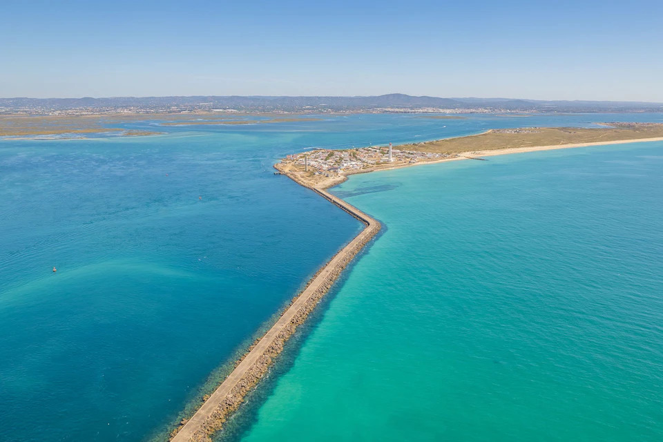 Aerial view of a long jetty leading to a coastal village with lighthouses, surrounded by turquoise and deep blue waters.