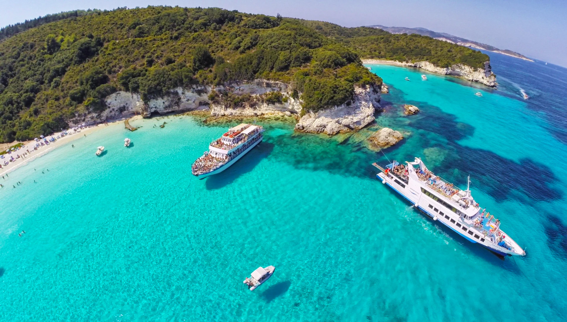 Turquoise waters with two large boats and a small boat near a rocky shoreline with lush greenery.