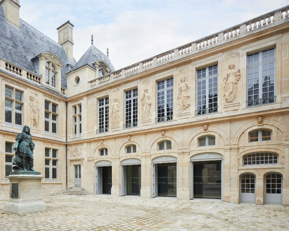 A historic building with ornate stone carvings, large arched windows, and a courtyard with a statue under a partly cloudy sky.