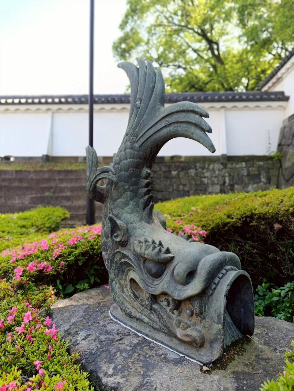 A close-up of a dragon-like sculpture outdoors, surrounded by greenery and pink flowers, with a white wall in the background.