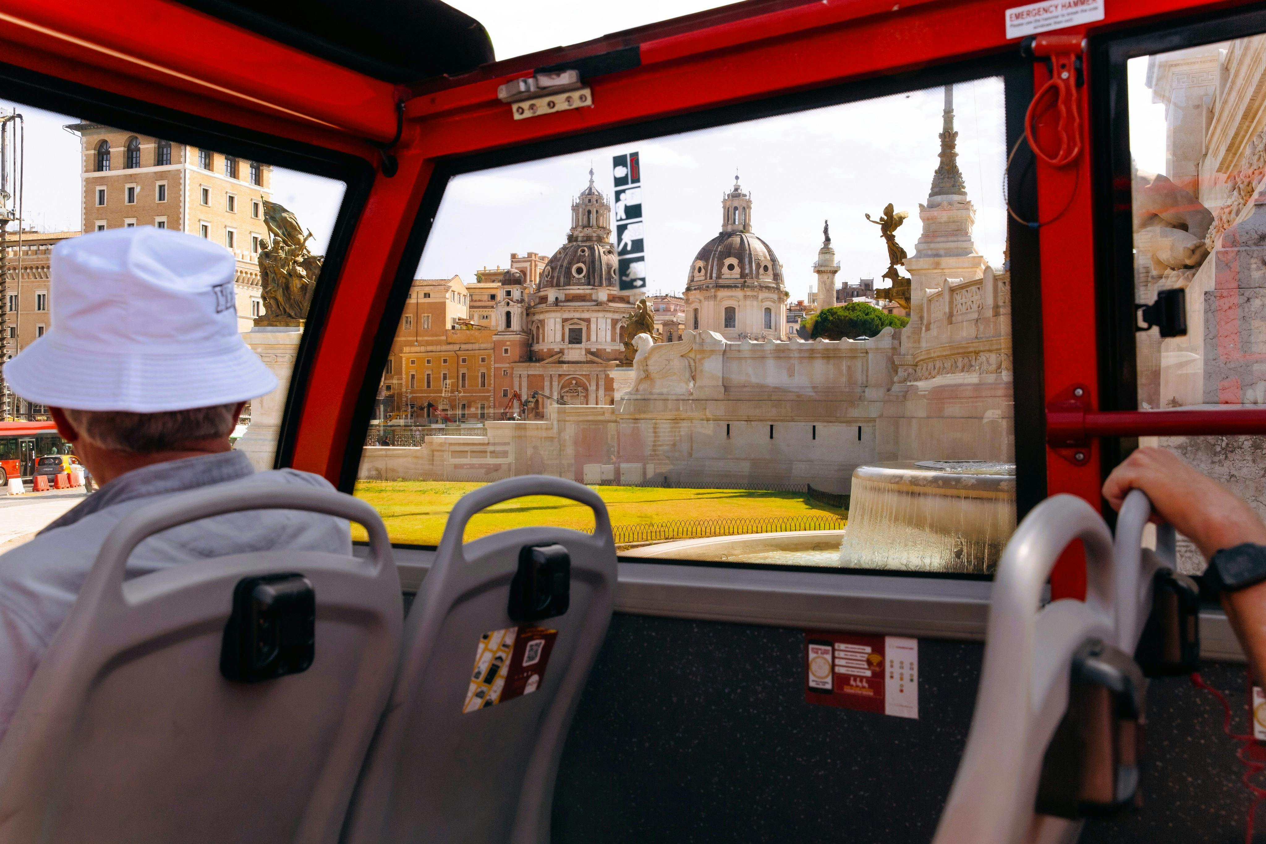 View of historic buildings and statues from inside a red bus with empty seats and a passenger wearing a white hat.
