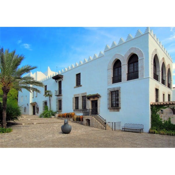 White, two-story building with arched windows, outdoor stairs, palm tree, and scattered pottery jars on a cobblestone pathway.
