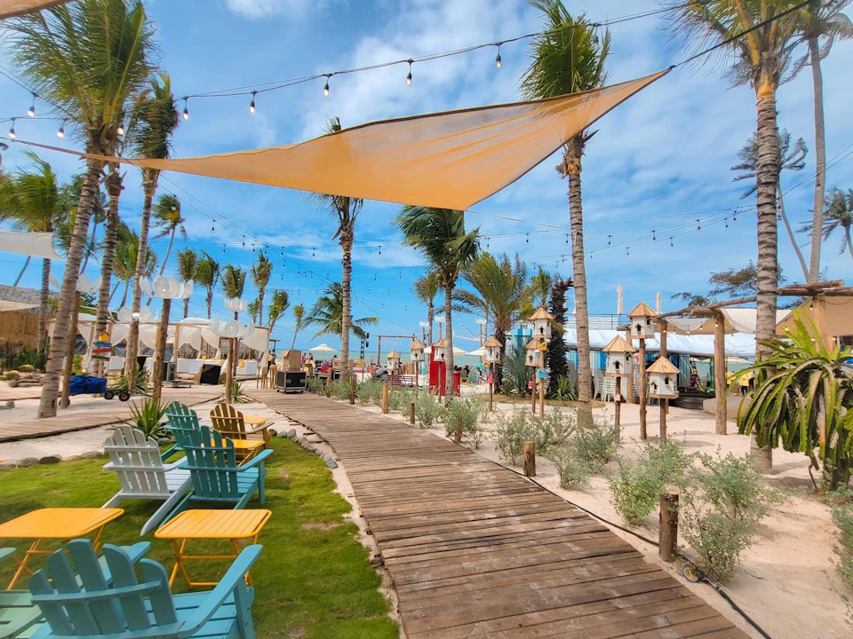 A beach area with a wooden path, palm trees, colorful chairs, hanging string lights, and a large fabric shade under a clear sky.