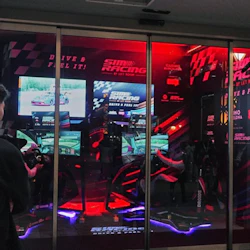 En grupp människor observerar och filmar en starkt upplyst sim racing setup med flera skärmar och racing simulator säten.