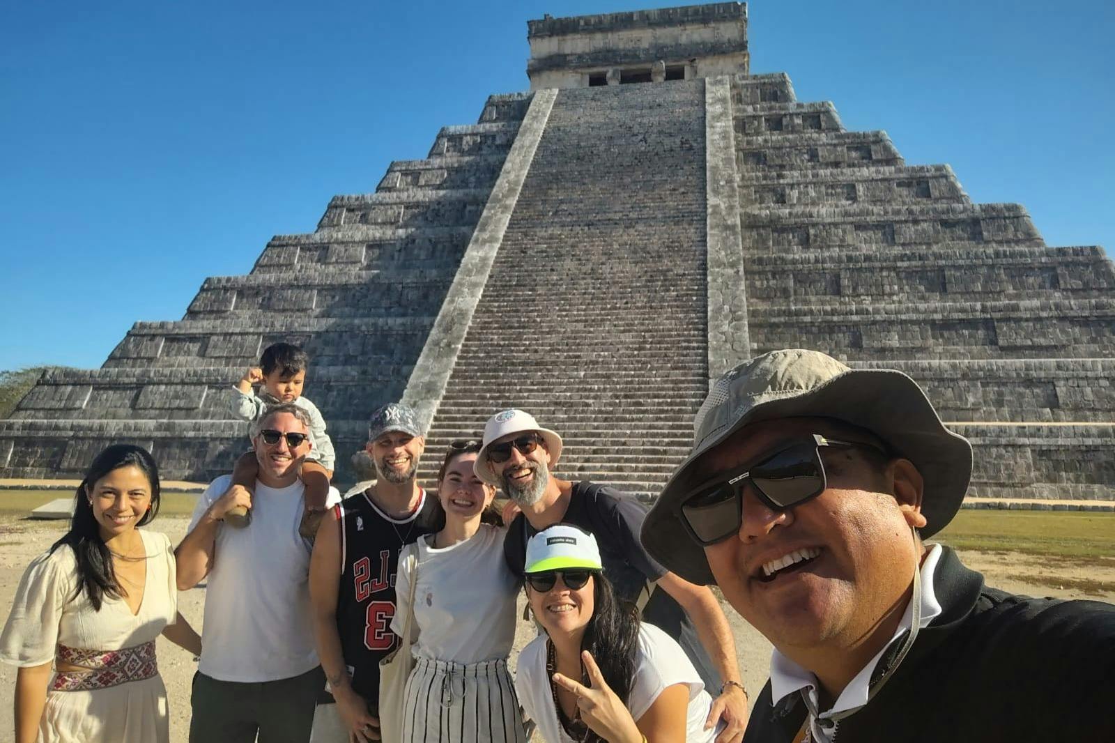Группа людей улыбается и позирует перед древней ступенчатой пирамидой под ясным голубым небом.
