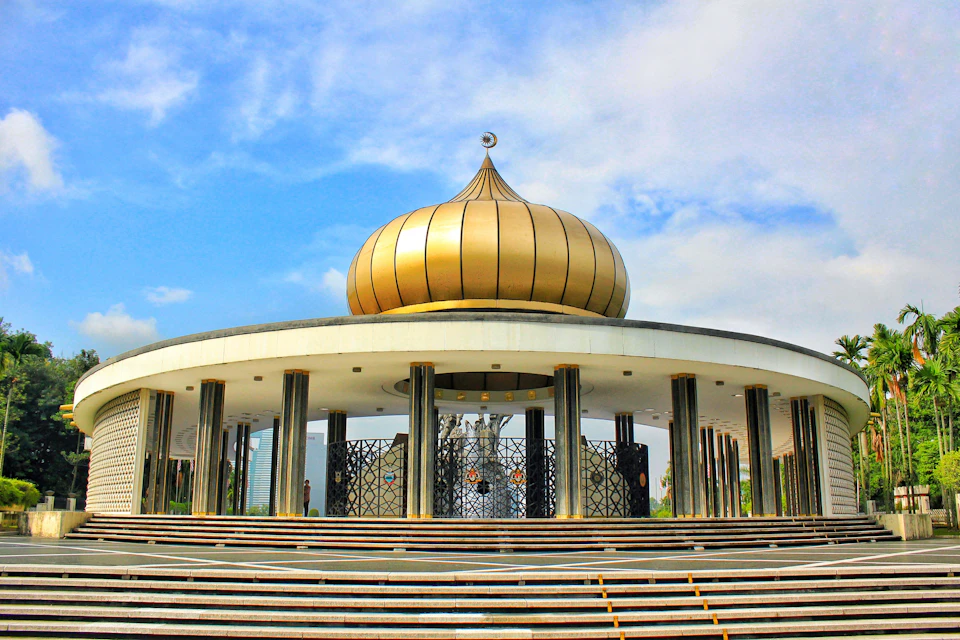 A monument with a large golden dome and black pillars, featuring circular steps leading up to it under a partly cloudy sky.