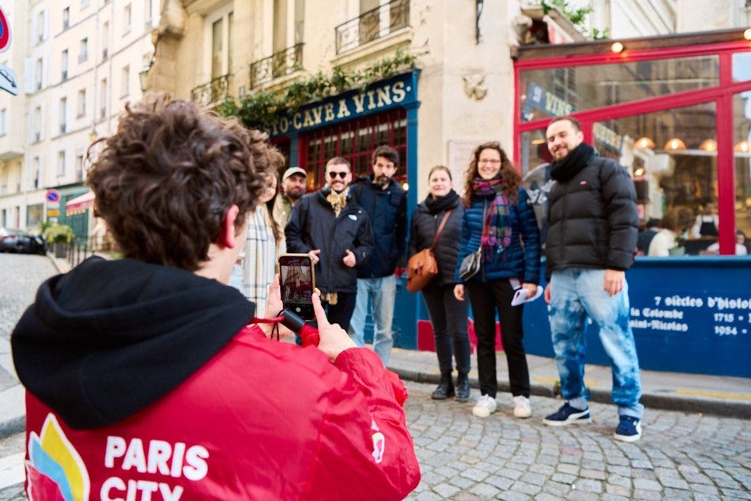 A person in a red jacket takes a photo of six people posing together in front of a blue storefront on a cobblestone street.