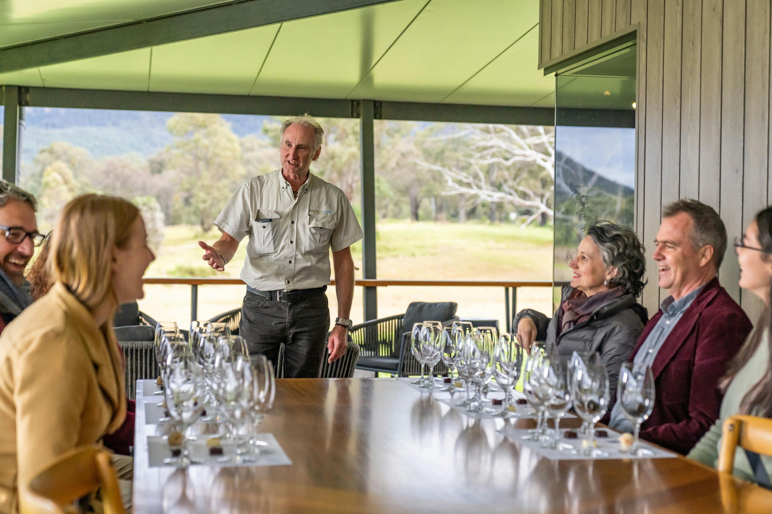 A man is speaking to a group seated around a table set with empty wine glasses in an outdoor venue with a scenic view.