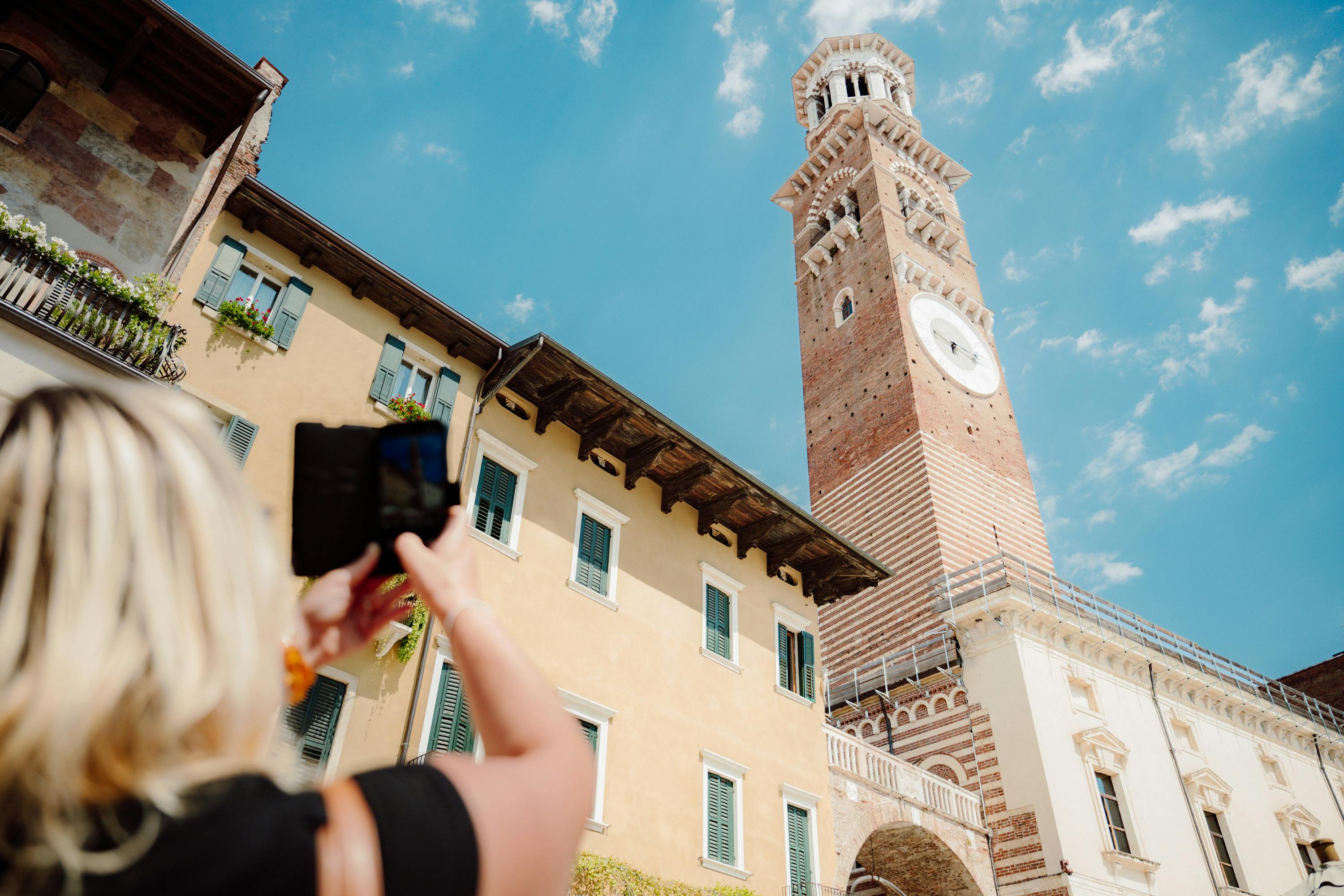 Una persona con in mano uno smartphone scatta una foto di un'alta torre dell'orologio accanto a un edificio sotto un cielo blu brillante.